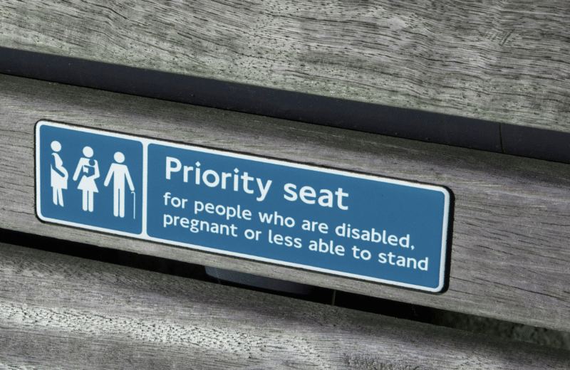 Tabliczka informacyjna z napisem „Priority seat for people who are disabled, pregnant or less able to stand”, umieszczona na drewnianej ławce – symbolicznie przedstawia uprzywilejowane miejsce siedzące dla osób z niepełnosprawnościami, kobiet w ciąży oraz osób z ograniczoną mobilnością.