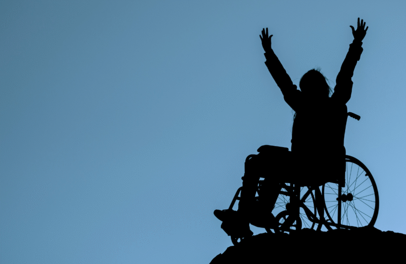 Osoba na wózku inwalidzkim z uniesionymi rękami na tle nieba – symbol determinacji i przełamywania ograniczeń.